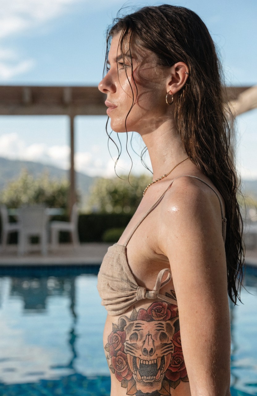 Woman in profile view displaying large tiger skull tattoo with roses on her side torso, next to a pool in a beige bikini top