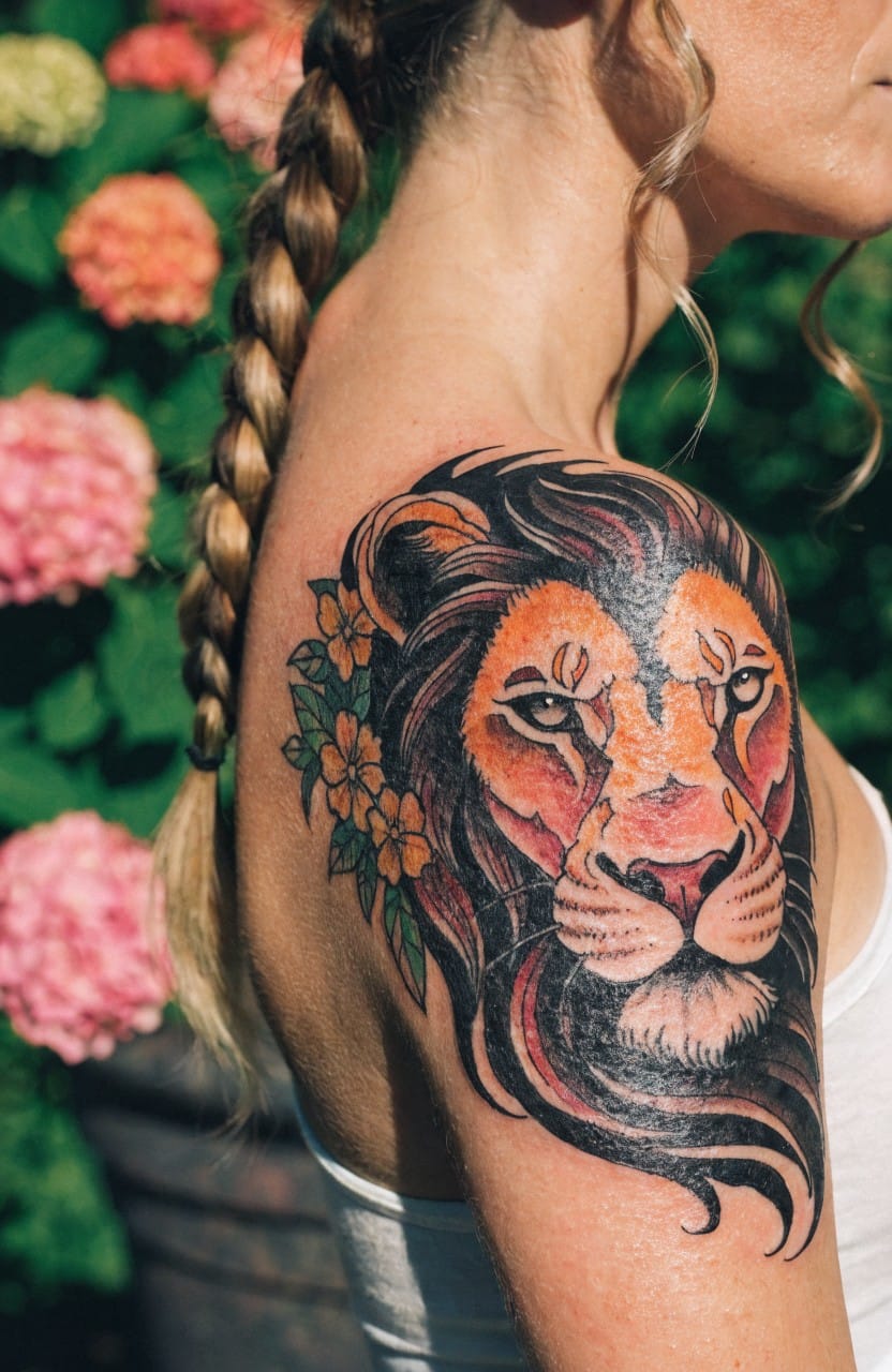 Woman's upper shoulder tattoo of a colorful lion head with yellow orange flowers in the mane against a garden backdrop