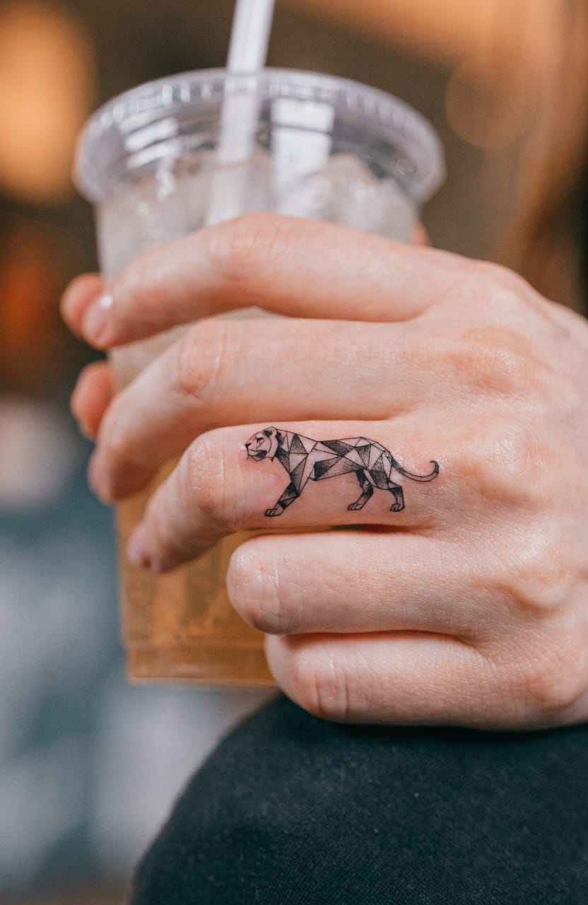 Hand holding a clear cup of iced drink with a small black geometric lioness tattoo on the ring finger