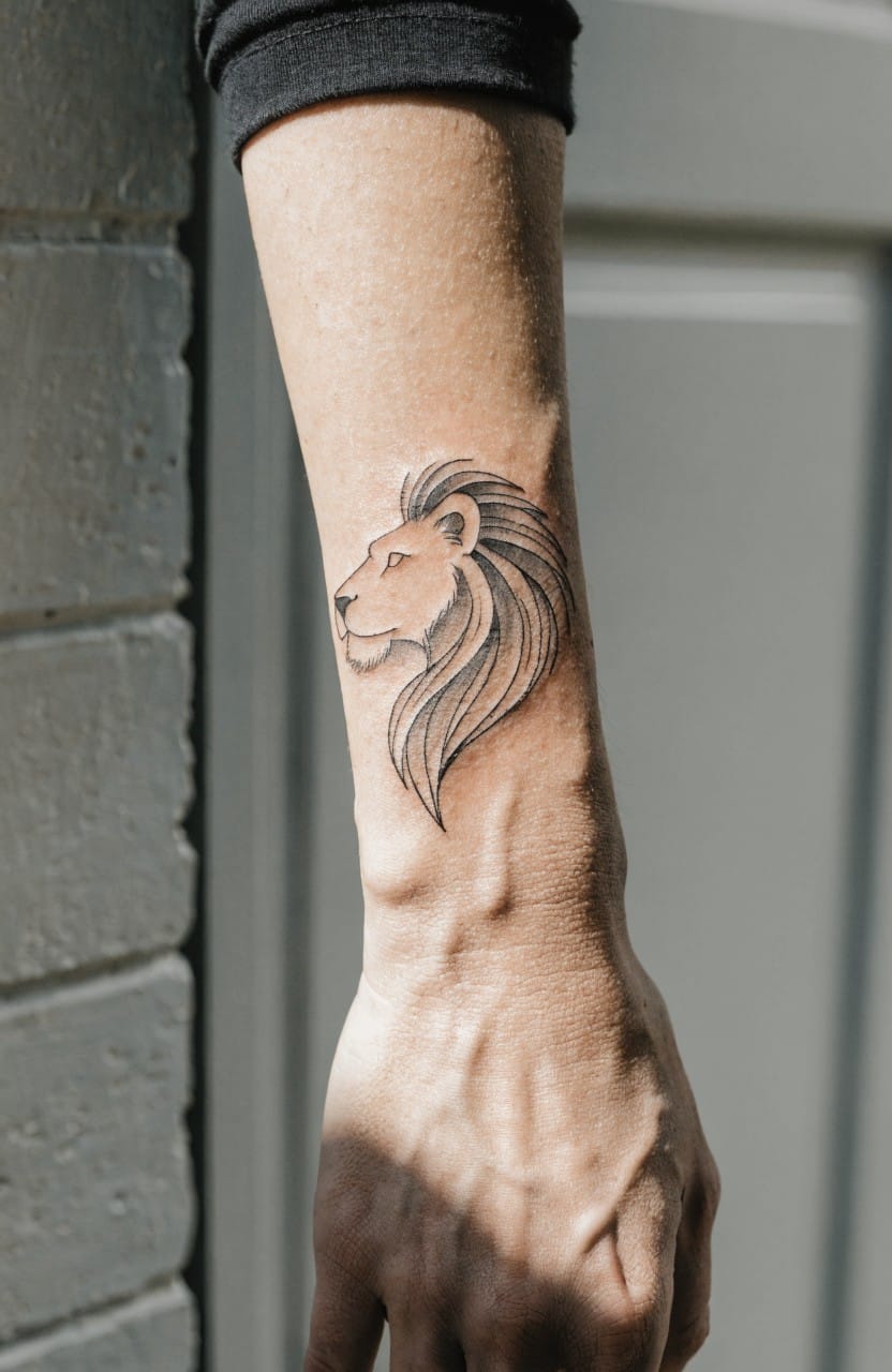 Black fine line tattoo of a lion head in profile on a forearm with flowing mane shading