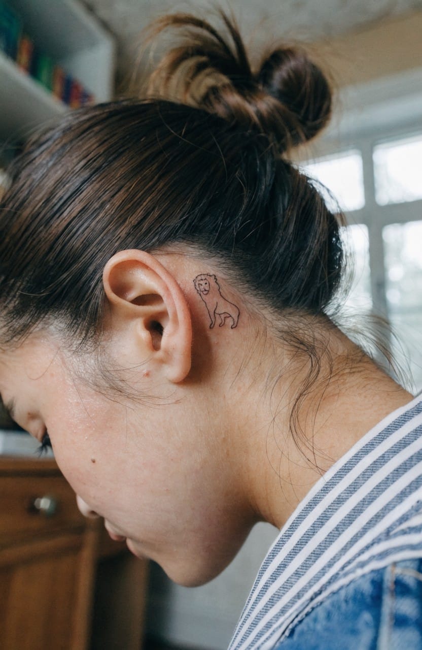 Woman's profile with hair in a bun revealing a small black line art lion tattoo behind her ear on the neck