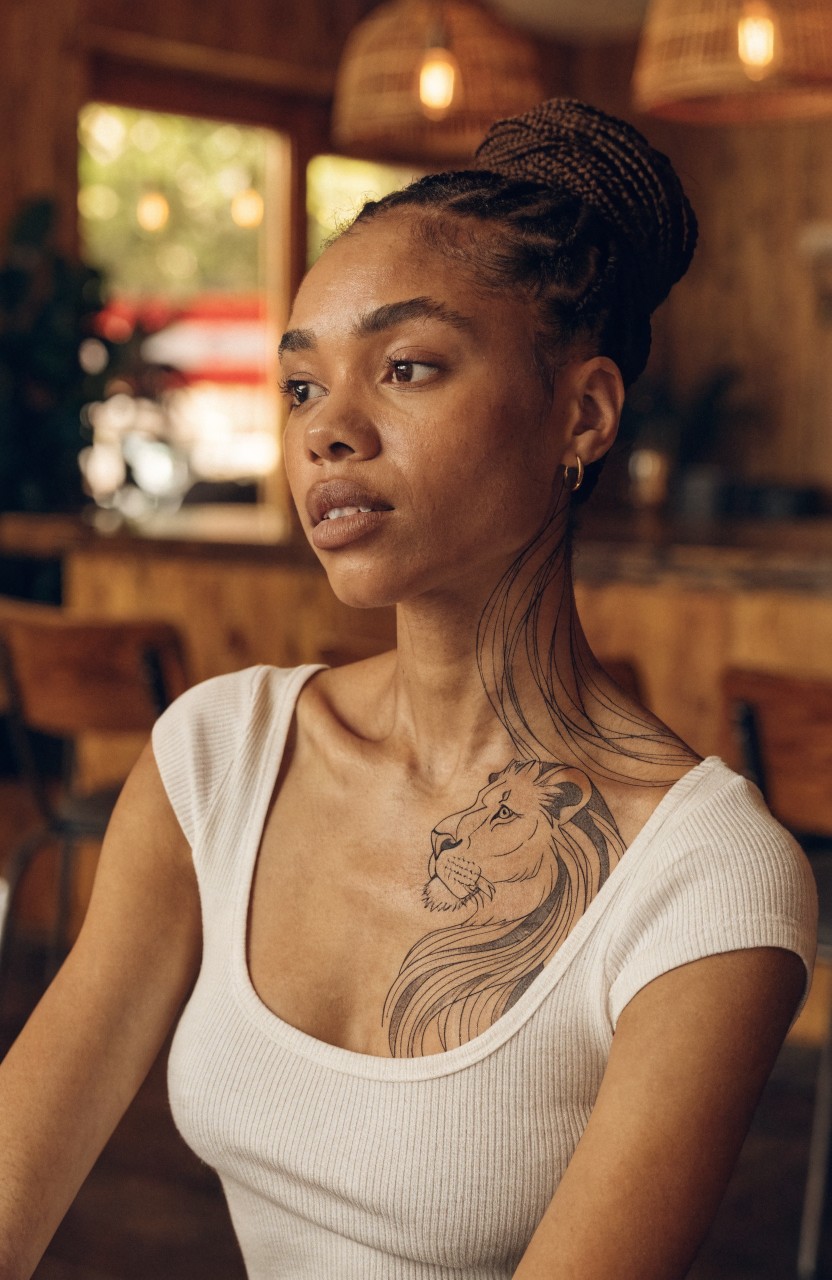 Black ink lion tattoo with flowing mane on a woman's chest and neck, visible in a white tank top against wooden bar stools