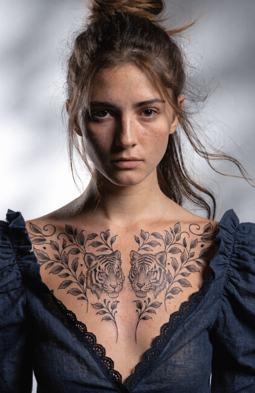 Symmetrical black ink tattoo of two facing tigers entwined with leaves on a woman's upper chest, shown in a low-cut navy blouse