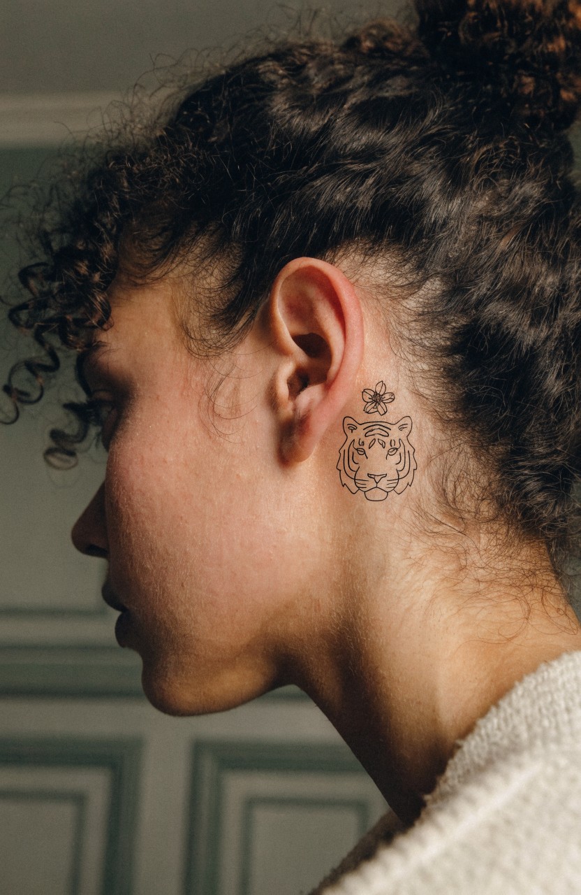 Profile view of a woman with a small black linework tiger head and flower tattoo behind her ear on the neck