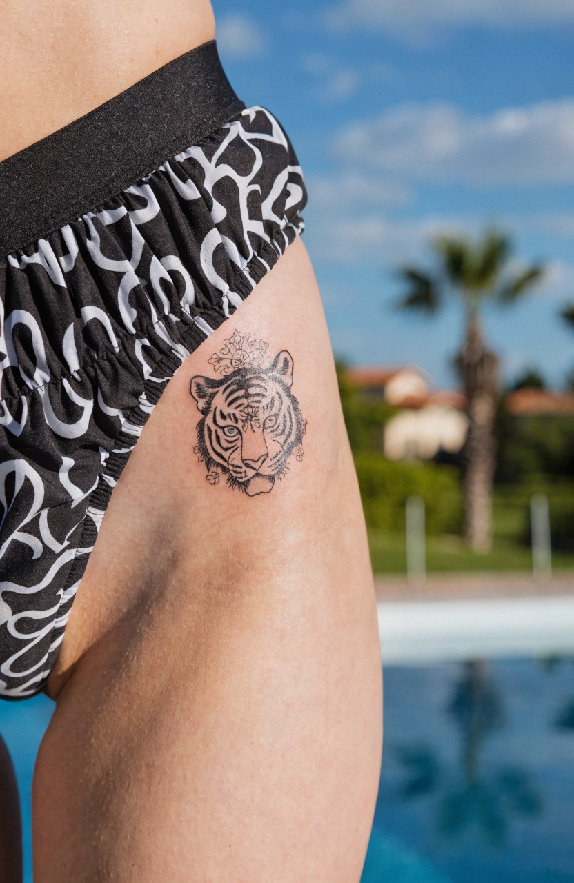 Black linework tiger head tattoo on upper thigh above bikini bottoms, detailed face with whiskers and eyes, poolside setting