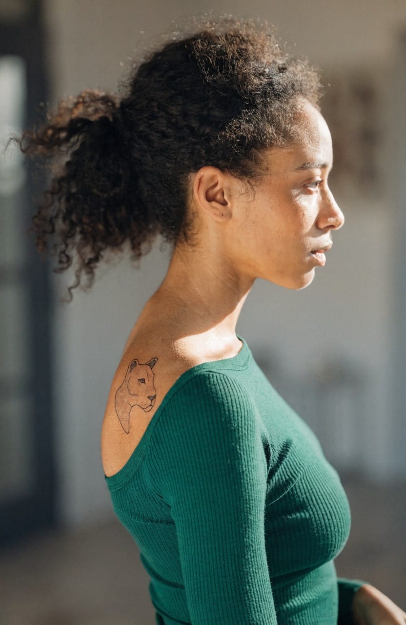 Minimalist black linework lioness head tattoo in profile on a woman's shoulder, peeking from an off-shoulder green top