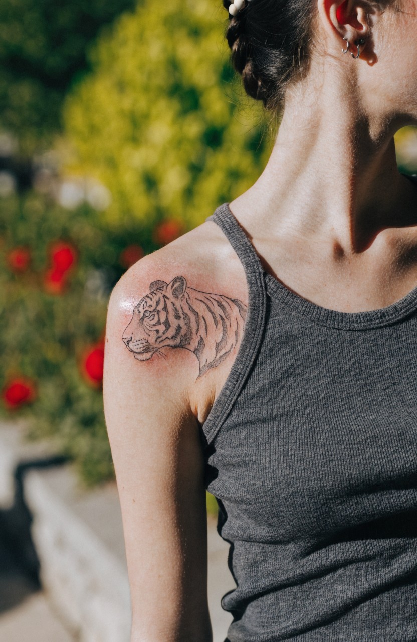 Woman's shoulder tattoo of a fine line tiger head in profile, shown on bare skin above a gray tank top strap, outdoors with green plants and red flowers in background