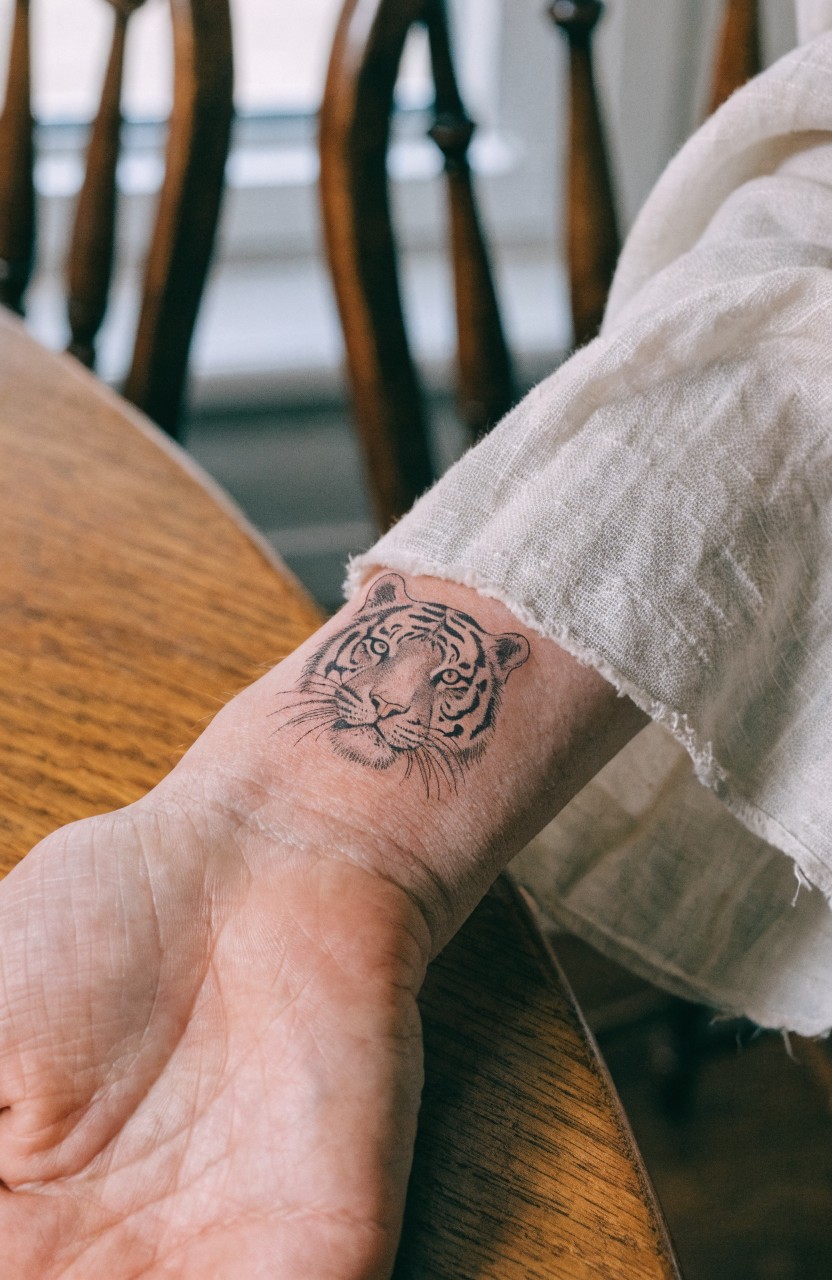 Fine line tiger head tattoo on inner forearm resting on wooden table with chairs and window in background