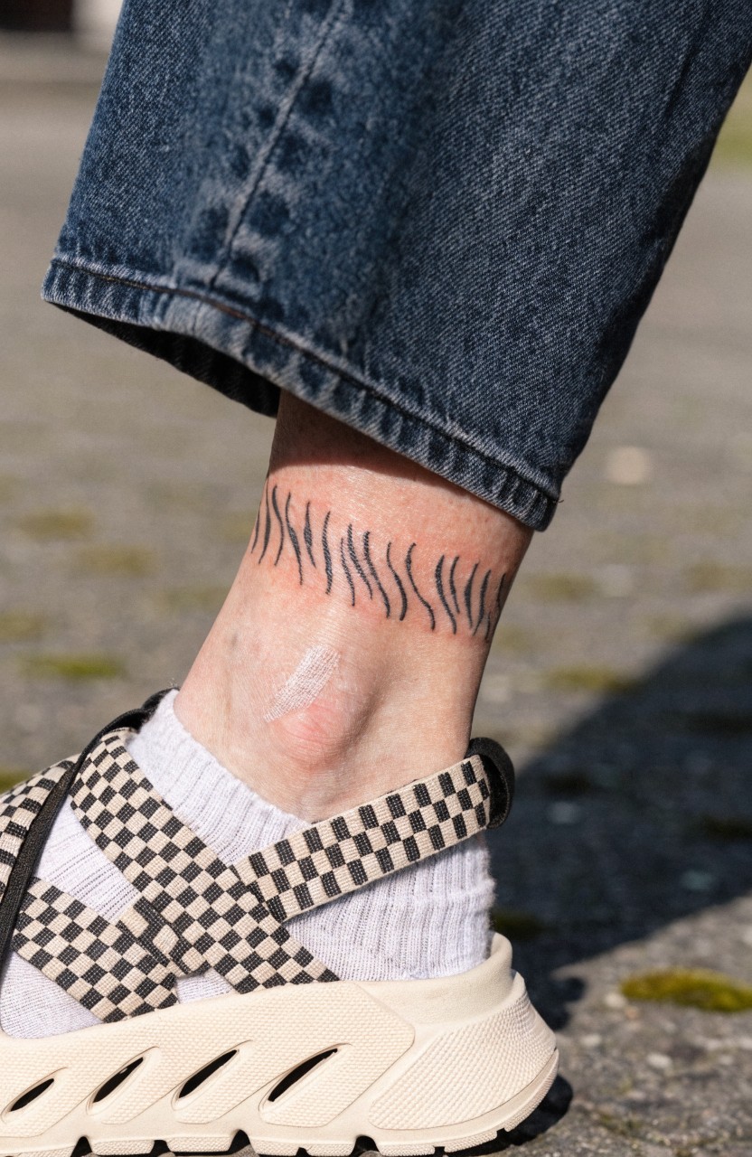 Close-up of a fine line tiger stripe tattoo wrapping around a person's ankle, inked in thin black lines on pale skin, paired with wide-leg jeans, checkered socks, and chunky sandals