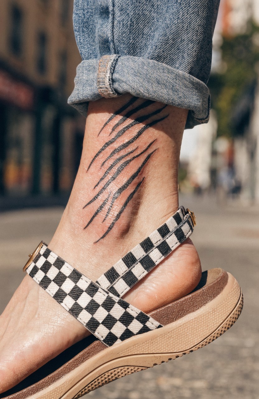 Fine line black tiger stripe tattoo wrapping around a woman's ankle, shown with rolled jeans and checkered sandal on a city street