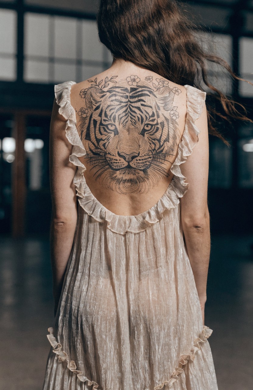 Back view of a woman in a sheer open-back dress revealing a large fine line tiger tattoo with floral accents on her upper back