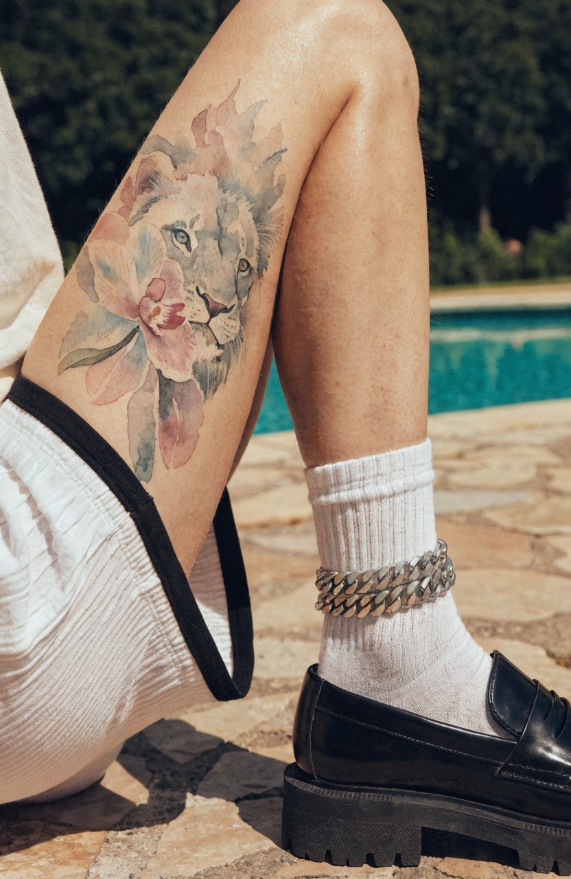 Colorful watercolor tattoo of lion head blended into pink flowers on thigh against poolside background