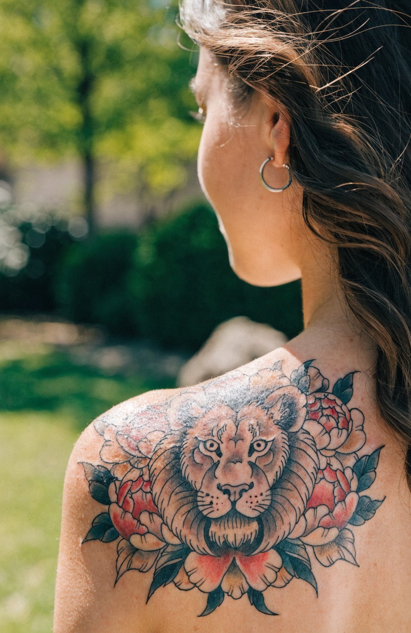 Colorful neo-traditional lion head tattoo blended with red and pink peony flowers on a woman's upper back and shoulder, photographed outdoors from behind