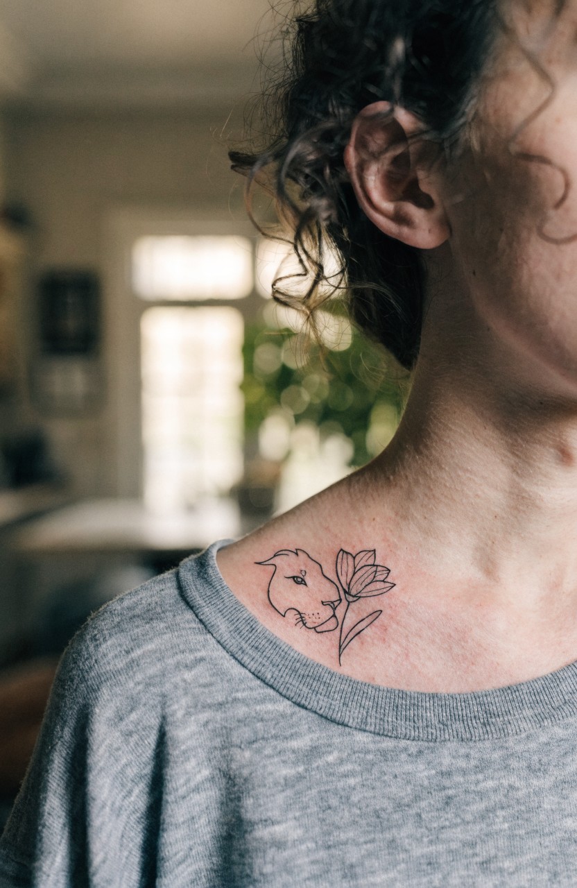 Black line art tattoo of lion face merging into tulip flower on woman's shoulder and collarbone