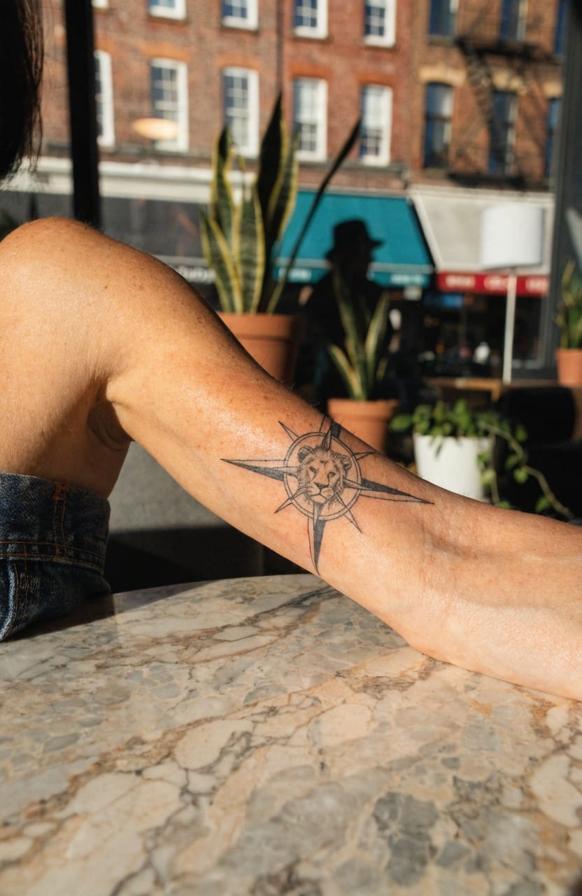 Black linework tattoo of lion head inside compass rose on woman's outer upper arm at outdoor cafe