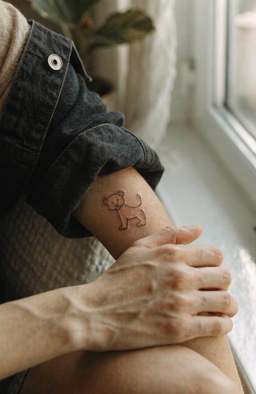 Minimalist black line art tattoo of a lion cub on a woman's inner forearm near the elbow, hand flexed nearby with natural light