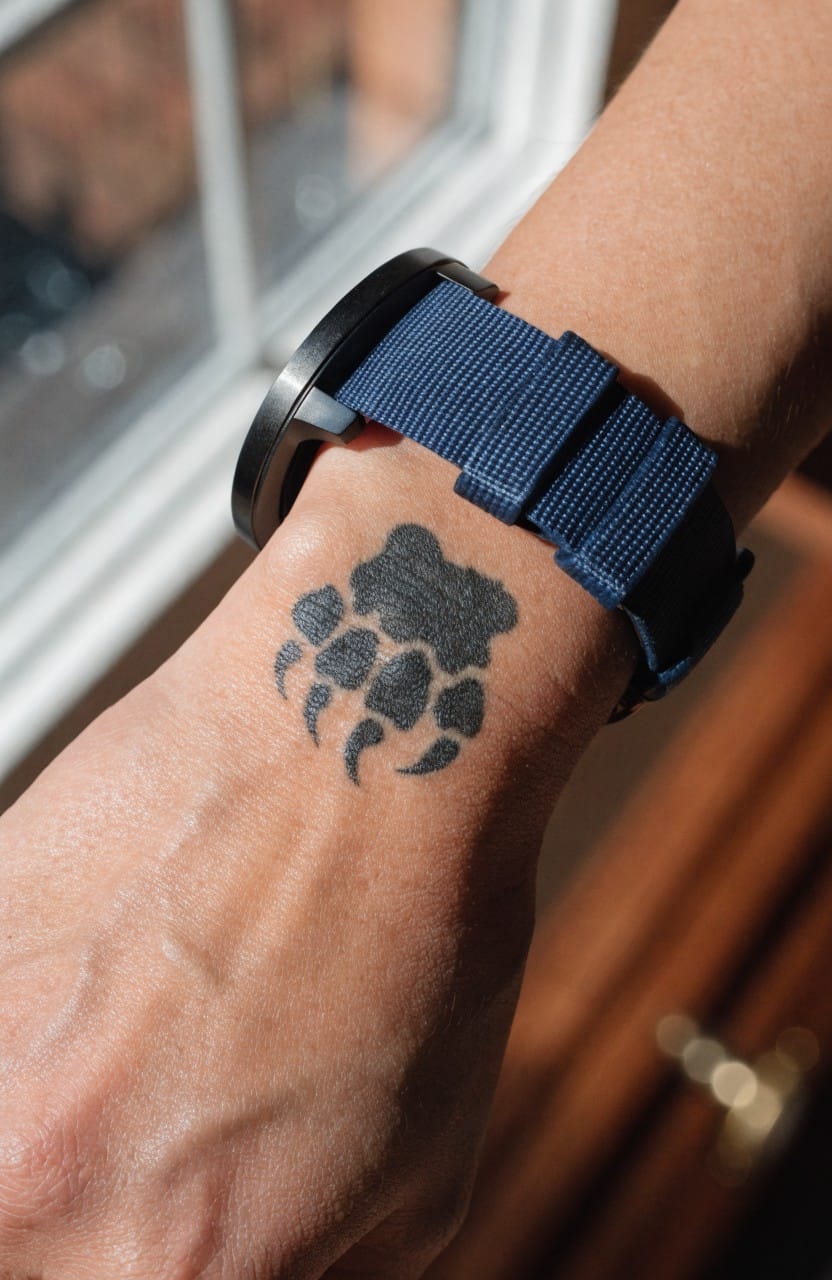 Black ink bear paw print tattoo on inner wrist next to a blue watch band