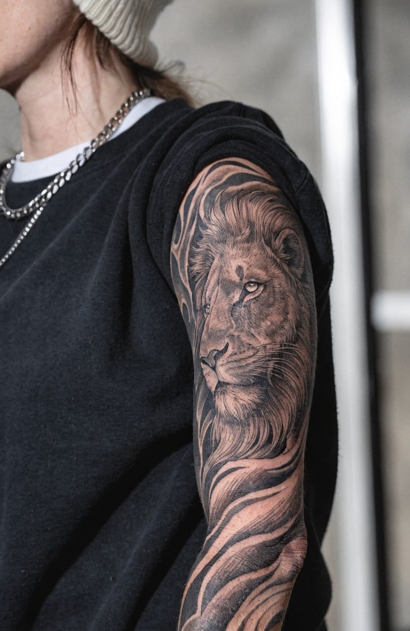 Woman's upper arm tattoo of a realistic black and grey lion head with flowing mane extending down the arm