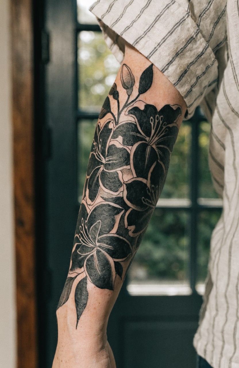 Black ink tiger lily tattoo wrapping fully around a forearm with shaded petals and trailing leaves, arm extended from rolled-up striped shirt near a window