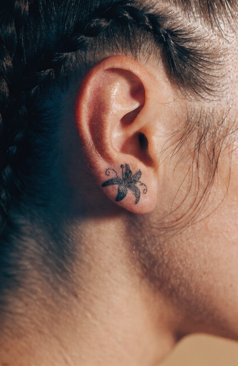 Small black ink tiger lily tattoo behind a woman's ear with subtle shading and braided hair framing it