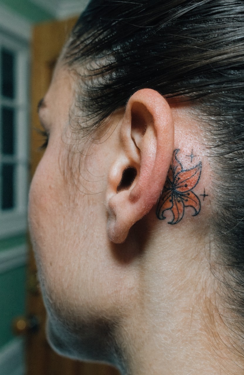 Small orange tiger lily tattoo with black outlines and stars behind a woman's ear on her neck