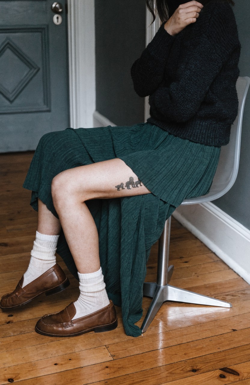 Black ink tattoo of a mama gorilla carrying her baby on her back, placed on a woman's upper thigh, visible under a green pleated skirt