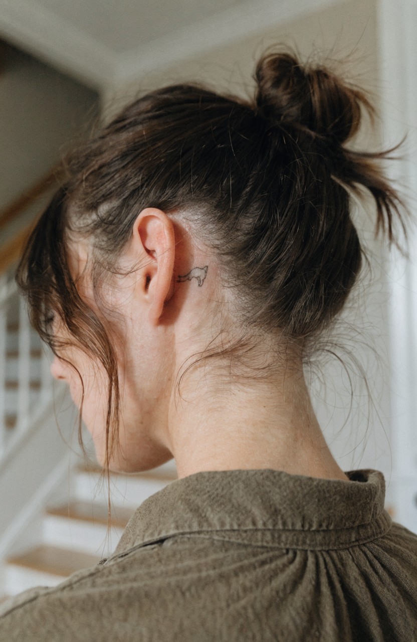 Small minimalist line tattoo of a lion cub behind a woman's ear on her scalp with hair pulled back