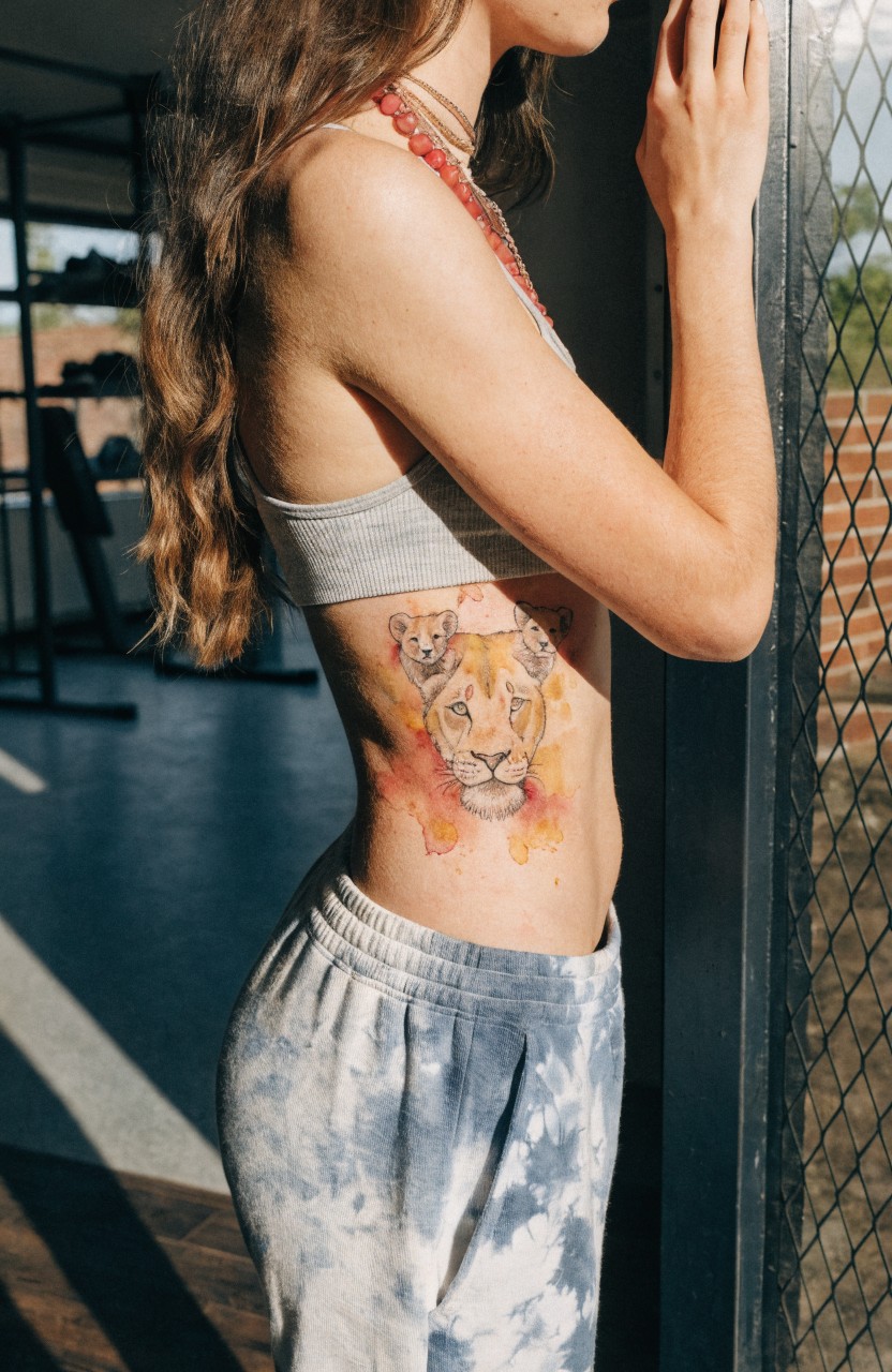 Woman showing watercolor lioness with two cubs tattoo on side torso in gym setting