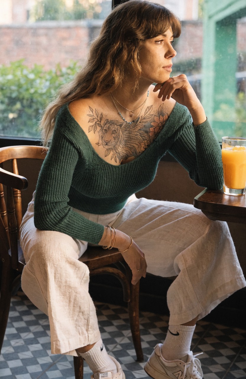 Black ink floral tattoo with shaded petals and leaves across a woman's upper chest, visible in a low-neck green sweater