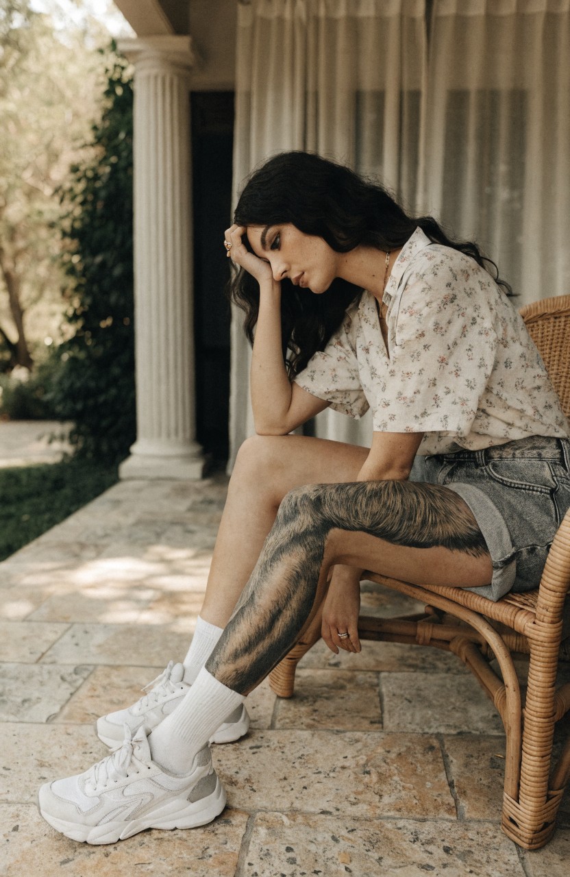 Woman's leg with a large black ink tiger sleeve tattoo wrapping from thigh to ankle, shown sitting on a porch chair wearing shorts and sneakers