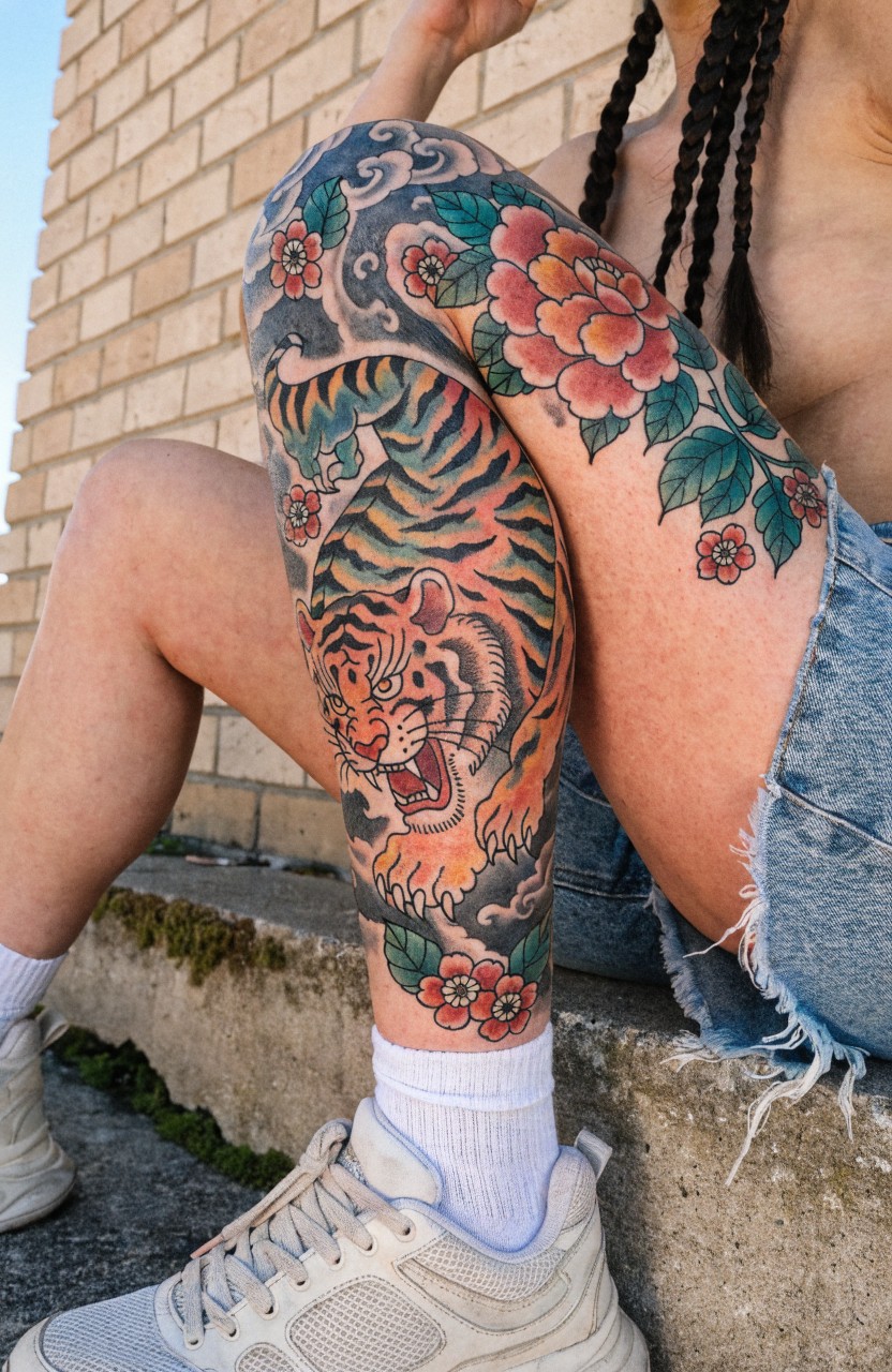 Woman's upper thigh tattooed with a vibrant Japanese-style tiger head and body integrated with red peonies and clouds, posed sitting on steps wearing shorts and sneakers