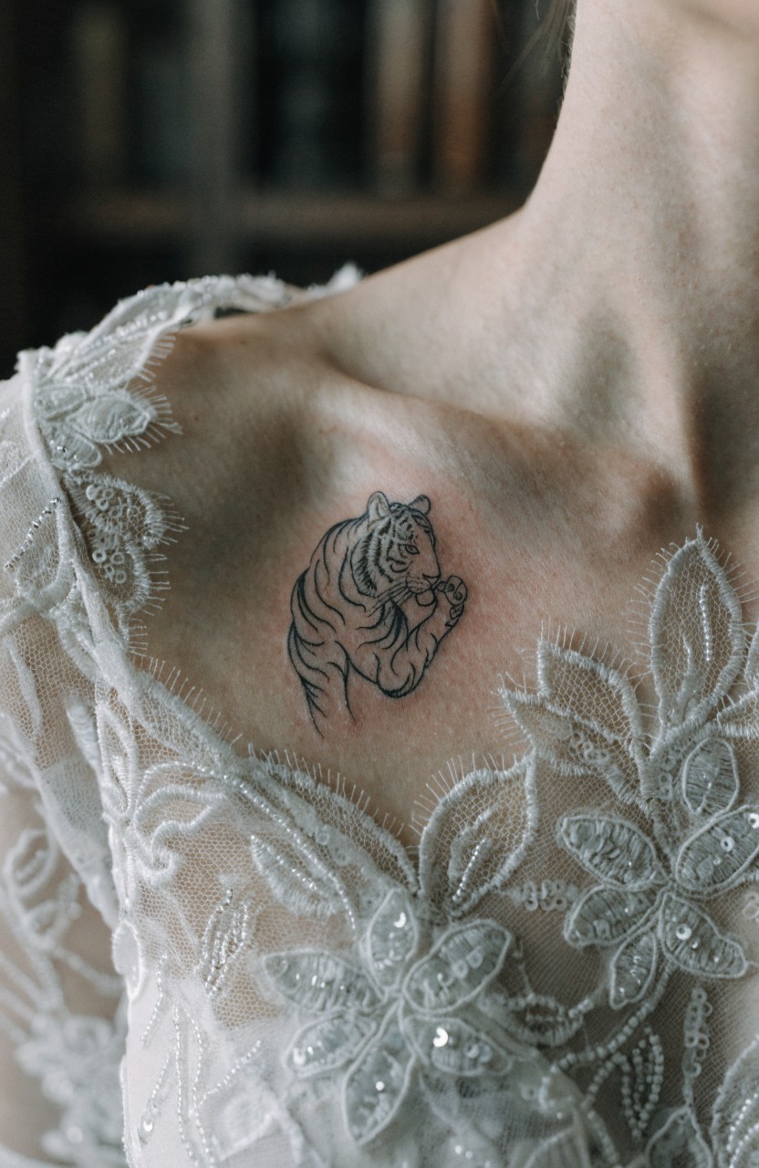 Small fine line tiger tattoo curled on a woman's collarbone under white lace dress fabric