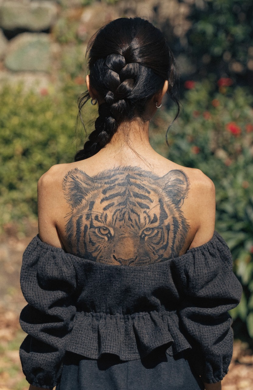 Woman viewed from behind with large realistic tiger head tattoo across upper back between shoulders, wearing off-shoulder black top in garden setting
