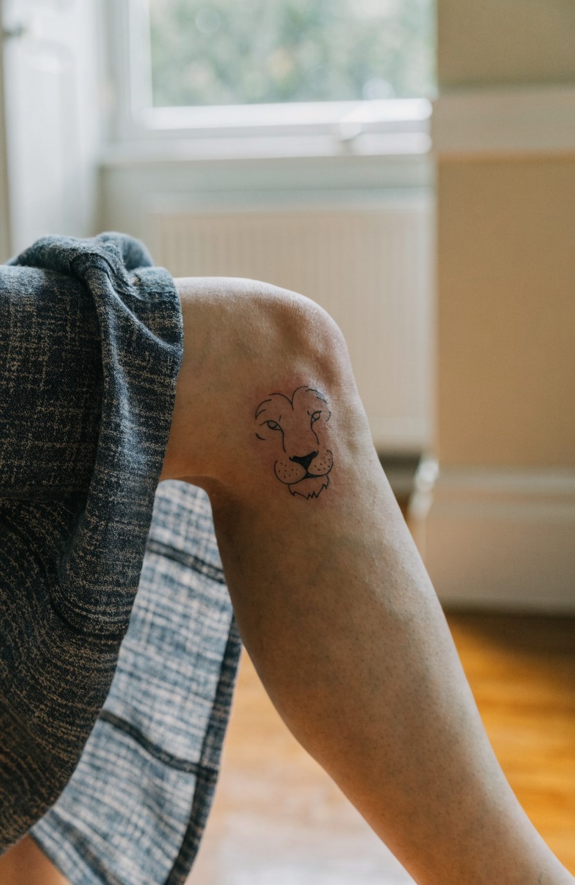 Small black linework lion face tattoo on a woman's knee with fine outlines subtle inner shading and textured gray fabric draped nearby