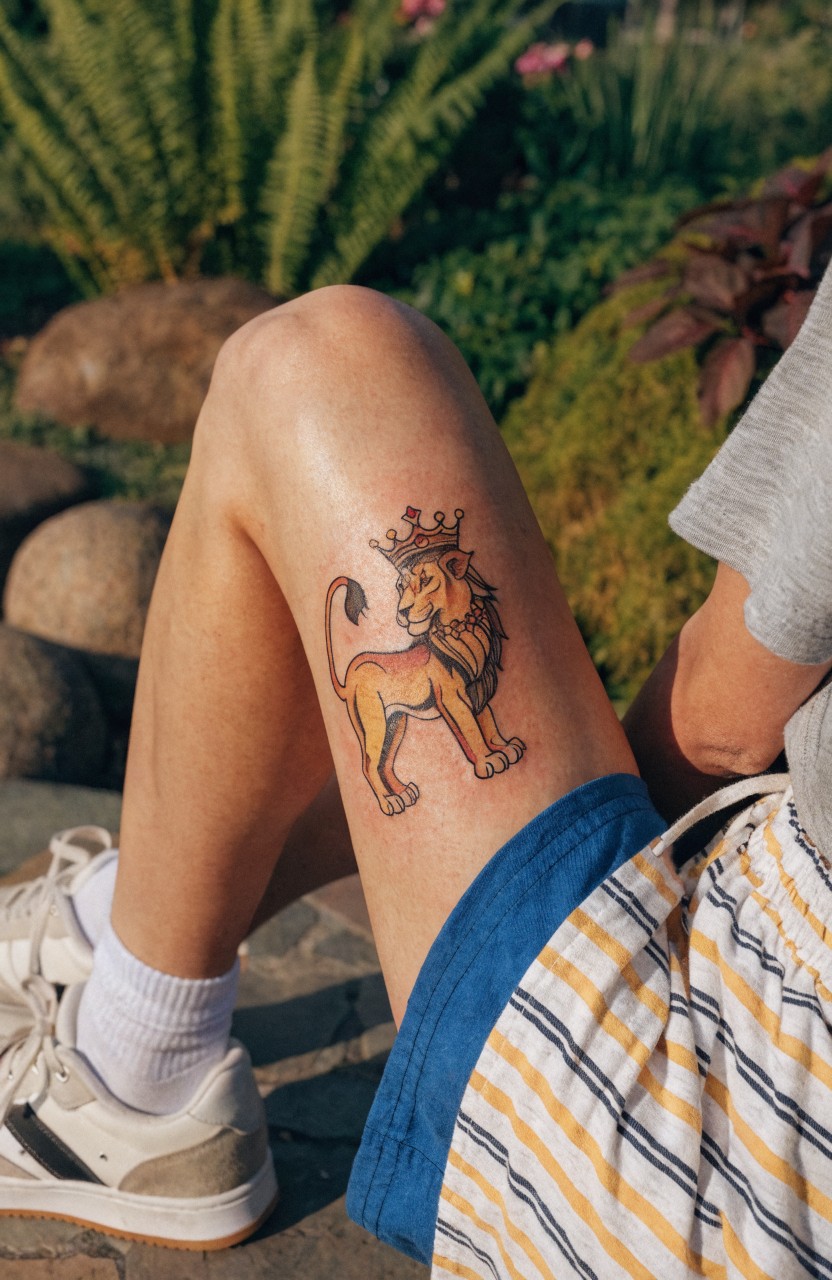 Dainty colorful lion with gold crown tattoo on upper outer thigh, leg posed on rocks amid garden plants