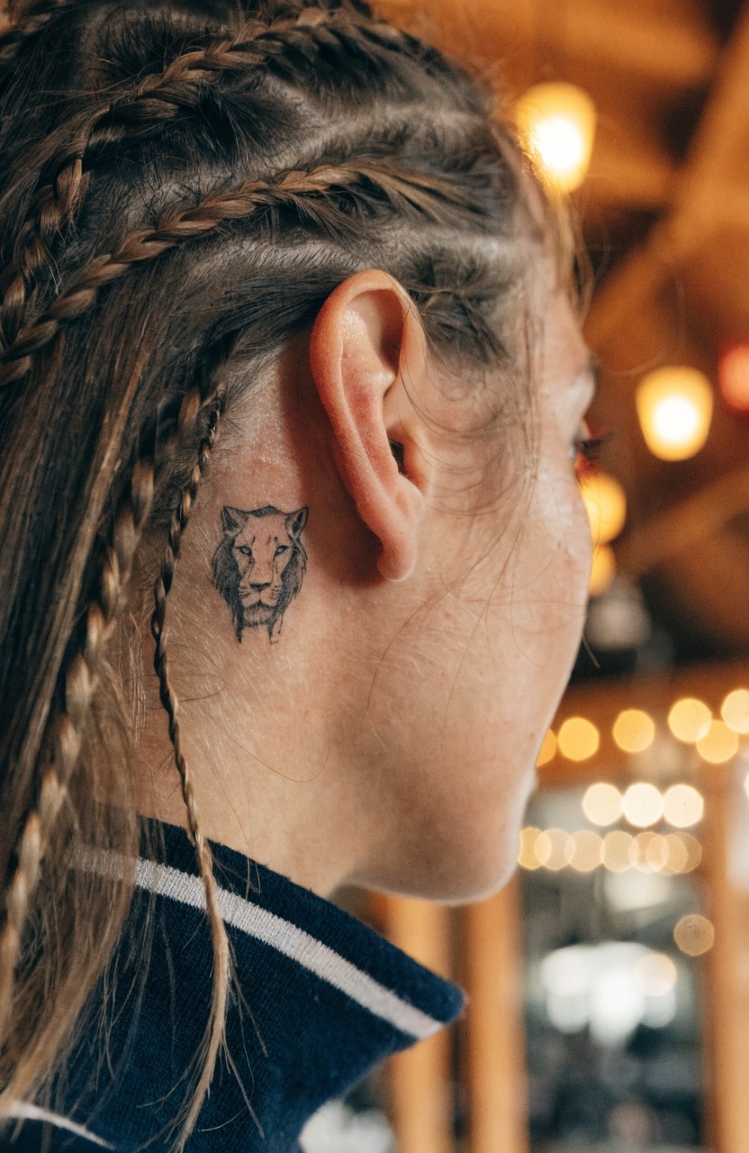 Woman's profile view with small black linework lion head tattoo behind her ear, framed by loose braids and warm interior lights