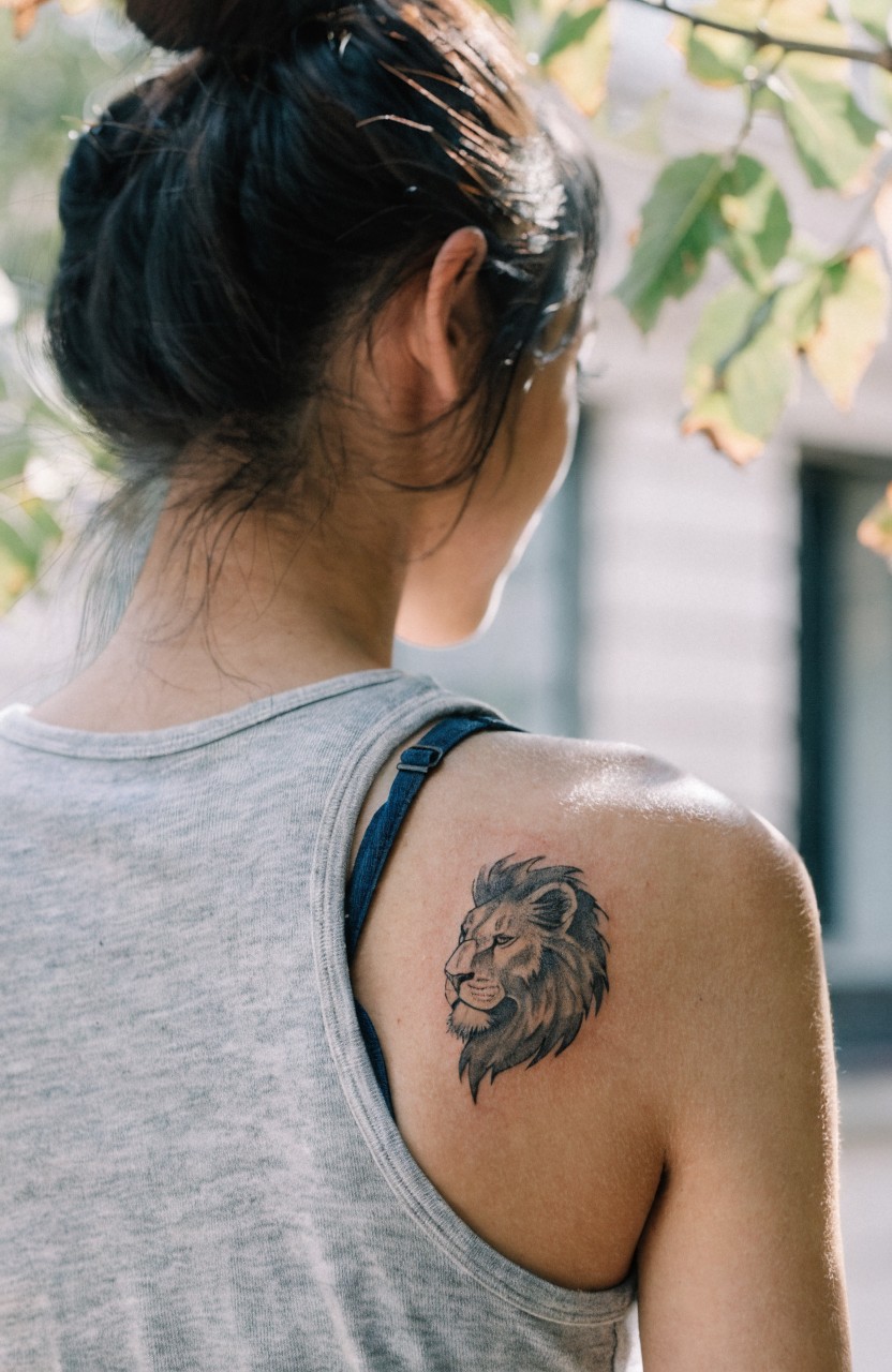 Small shaded black lion head tattoo on a woman's shoulder in a tank top, viewed from behind