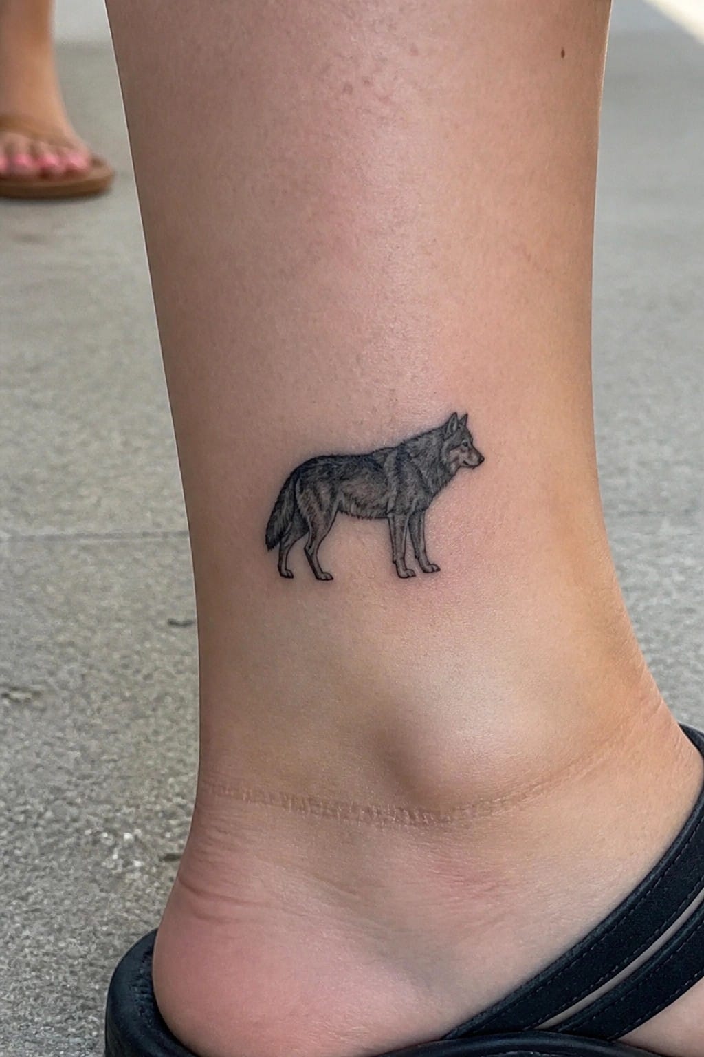 Wolf tattoo on ankle above black sandal, pink toenails visible in background