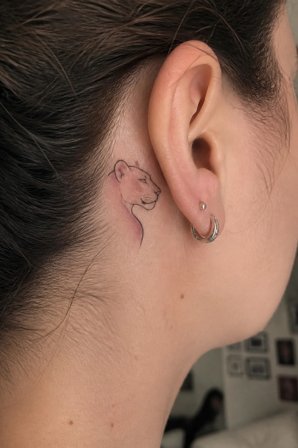 Small watercolor lioness tattoo in profile placed behind a woman's ear with soft pink tones and thin black outlines