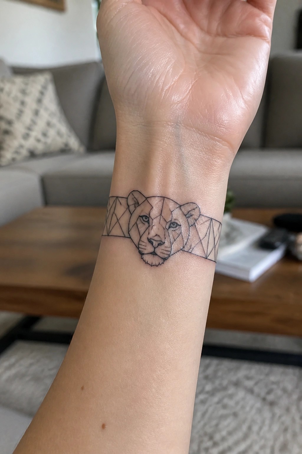 Geometric lion tattoo on wrist with blurred living room background visible behind.