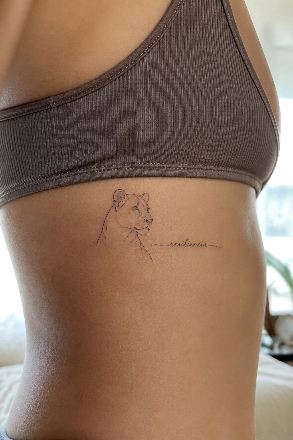 Fine line lioness head profile tattoo on the ribs with the word resiliencia written beside it in script.