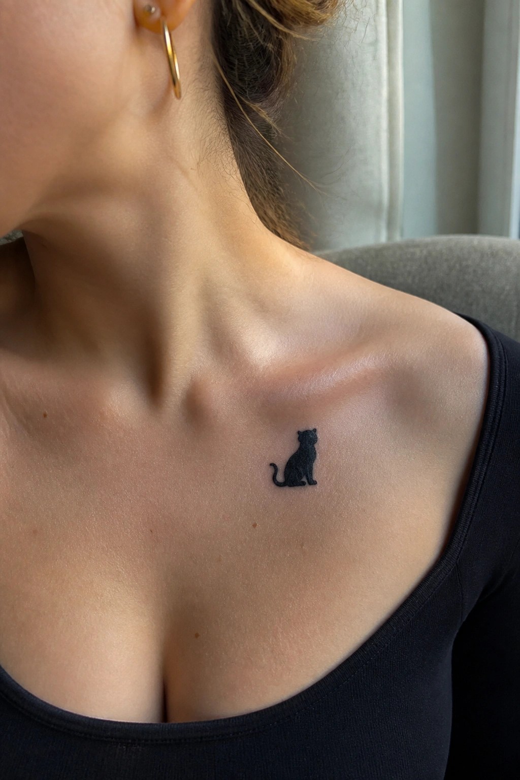 Small black cat tattoo on woman's collarbone above black top with gold earring