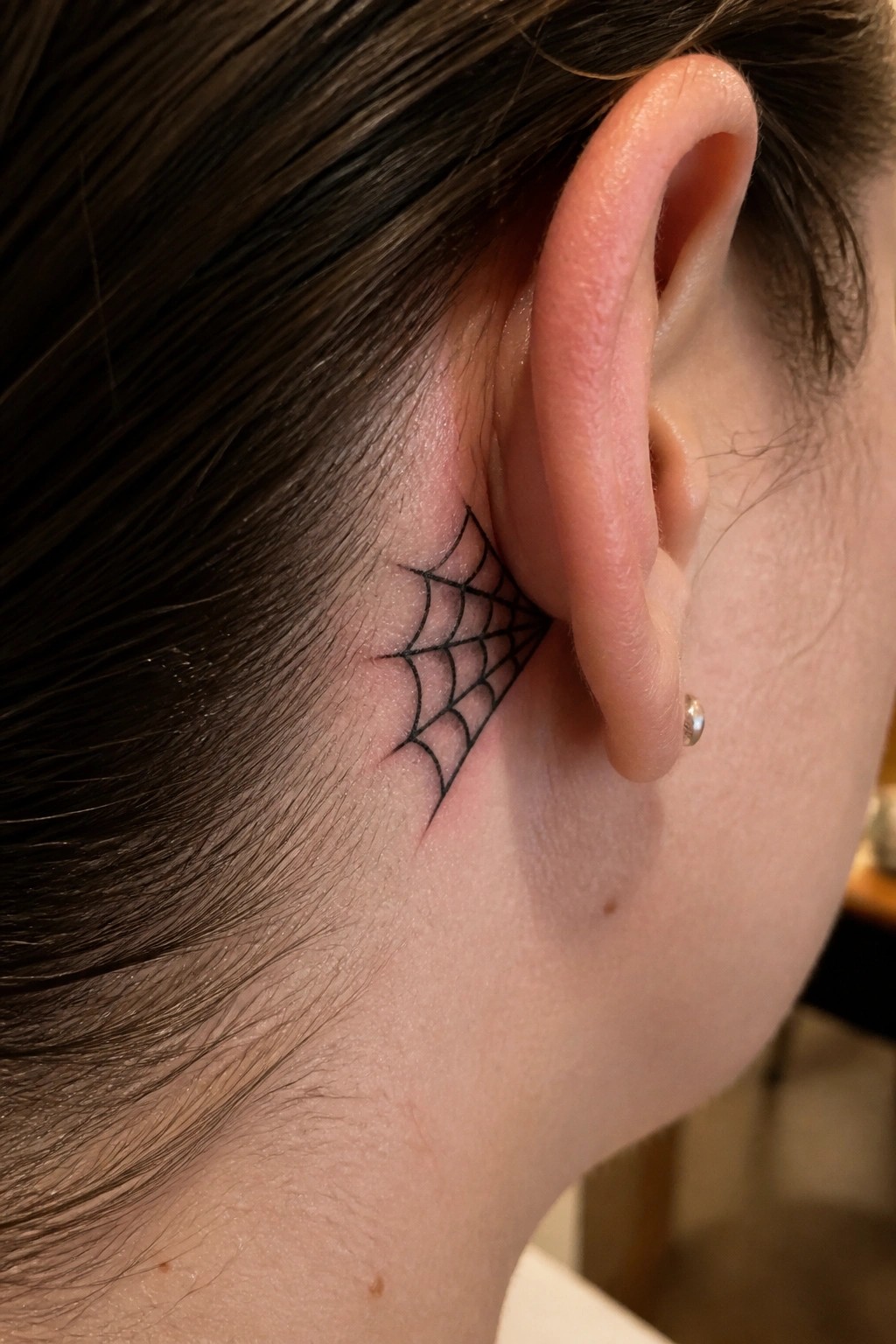 Black spiderweb tattoo behind ear with dark hair and small stud earring.