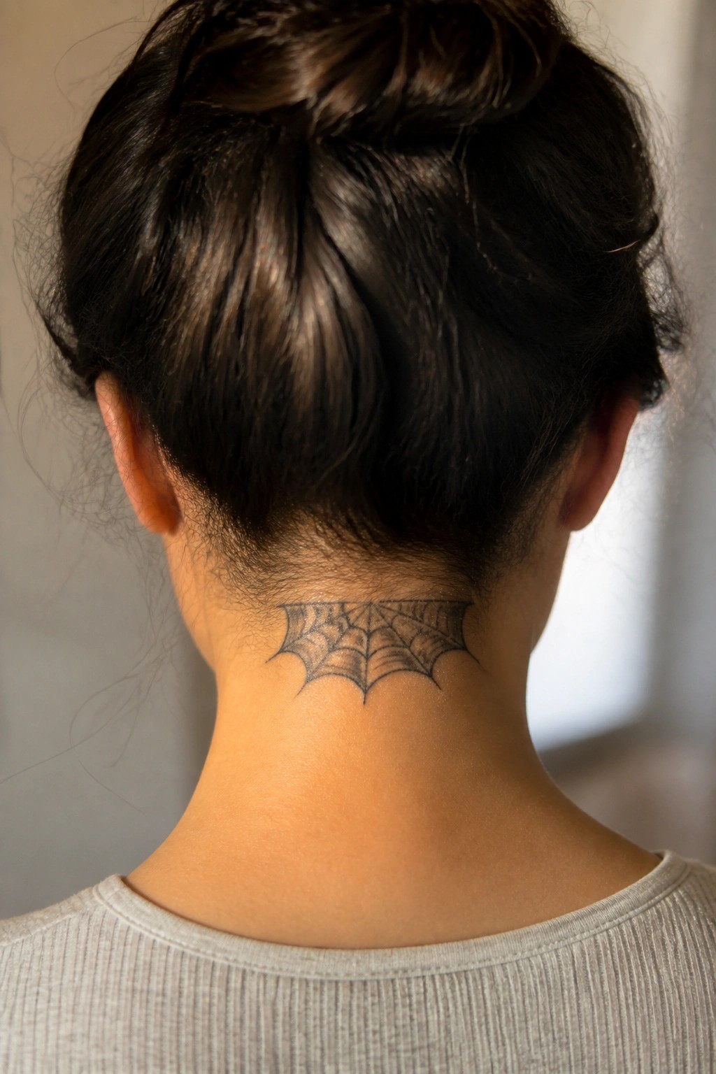 Dark-haired person in gray shirt with spiderweb tattoo on nape of neck