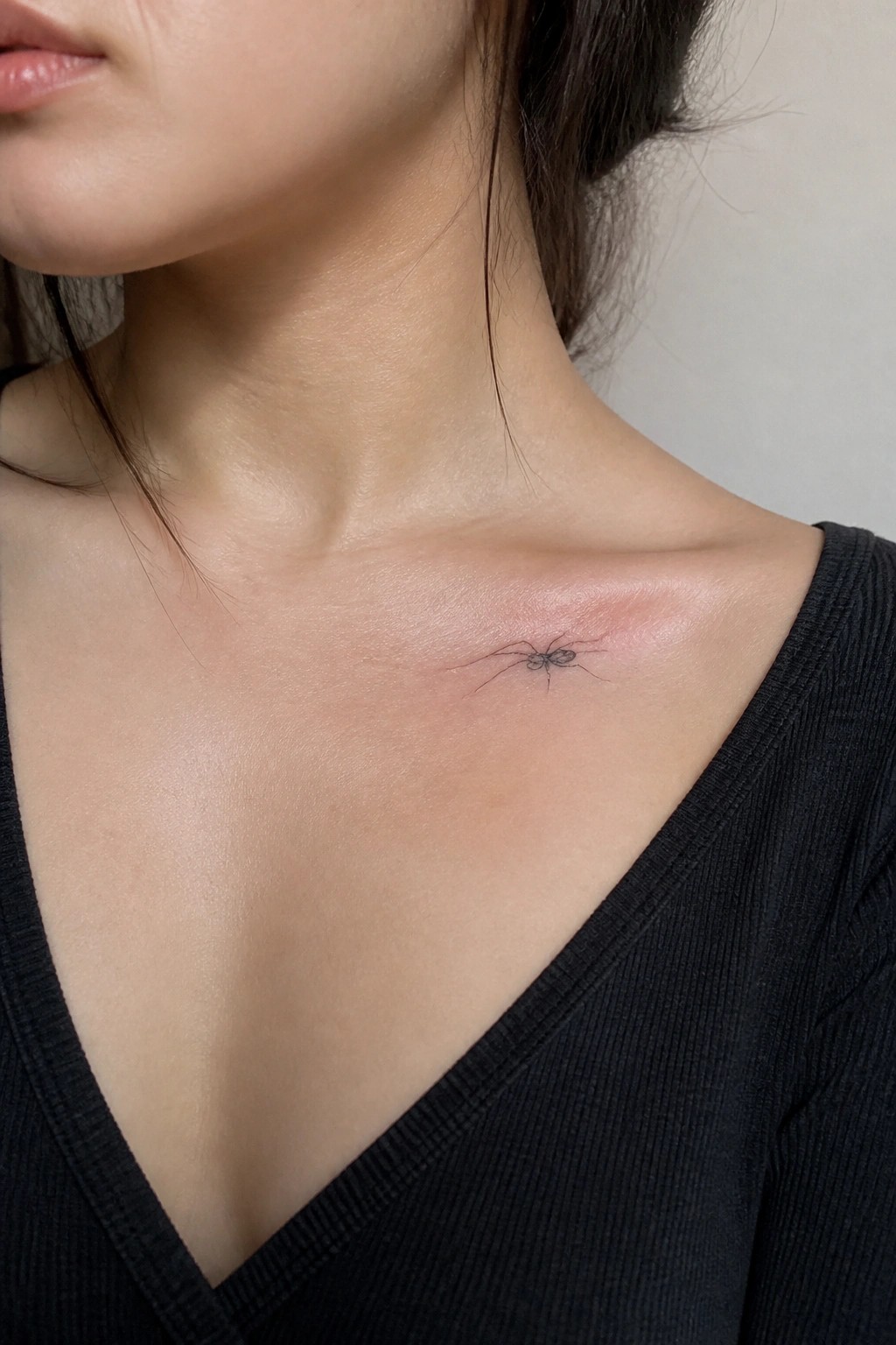 Small line art spider tattoo placed on the collarbone of a woman wearing a black top.
