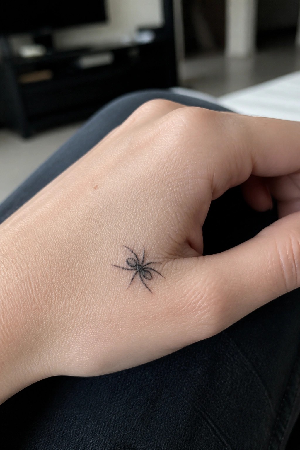 Hand with small black spider tattoo resting on dark jeans.