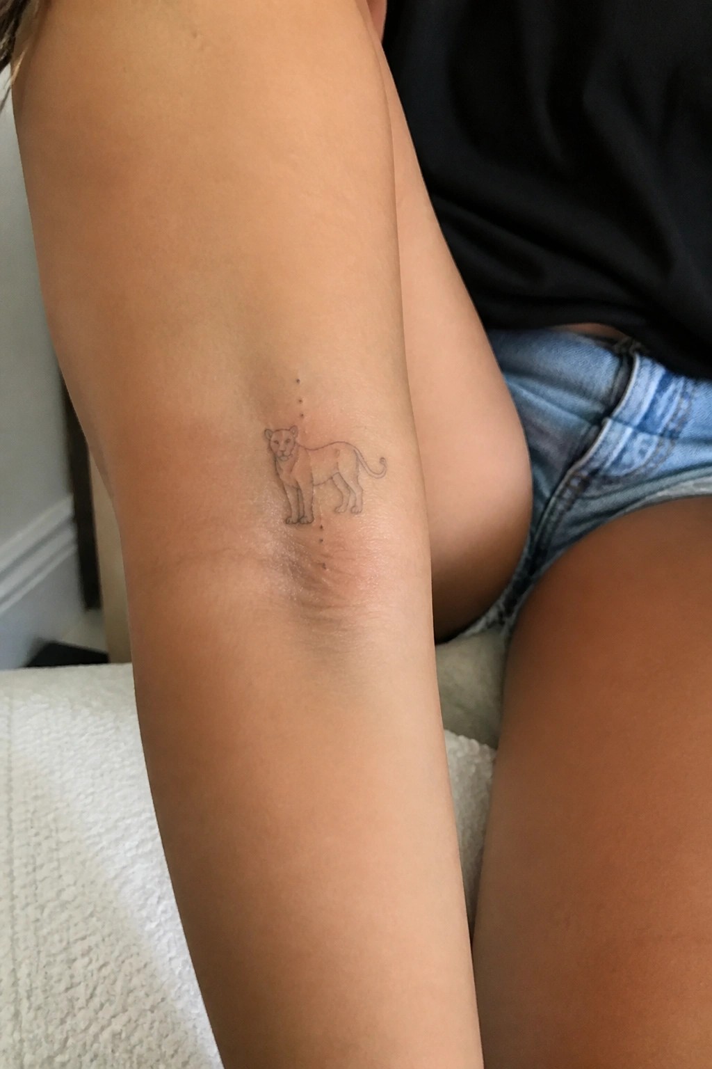 Minimalist lioness tattoo with dotted lines on upper arm beside denim shorts