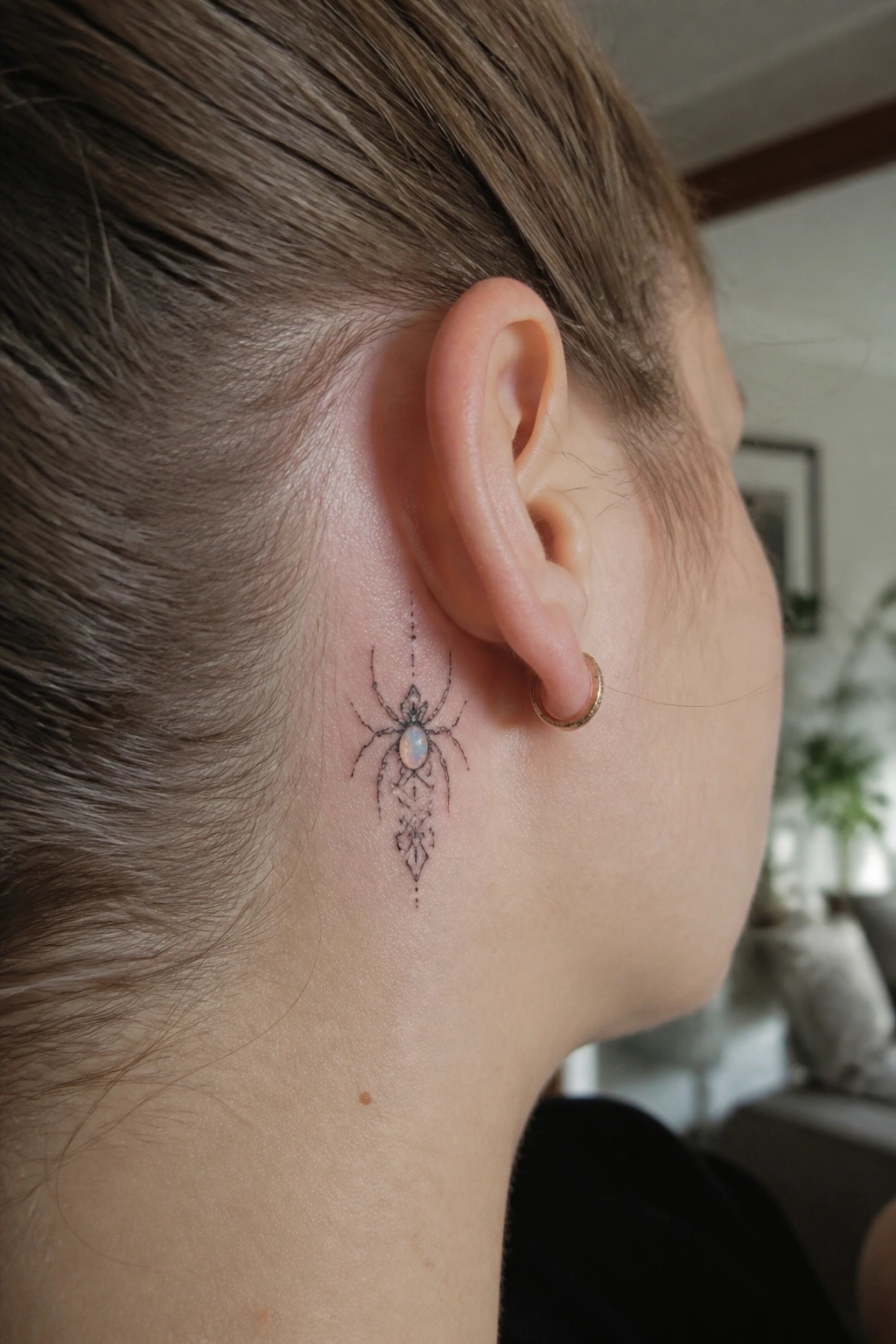 Small symmetrical spider tattoo with a central gem placed behind a woman's ear, featuring fine line ornamental patterns.