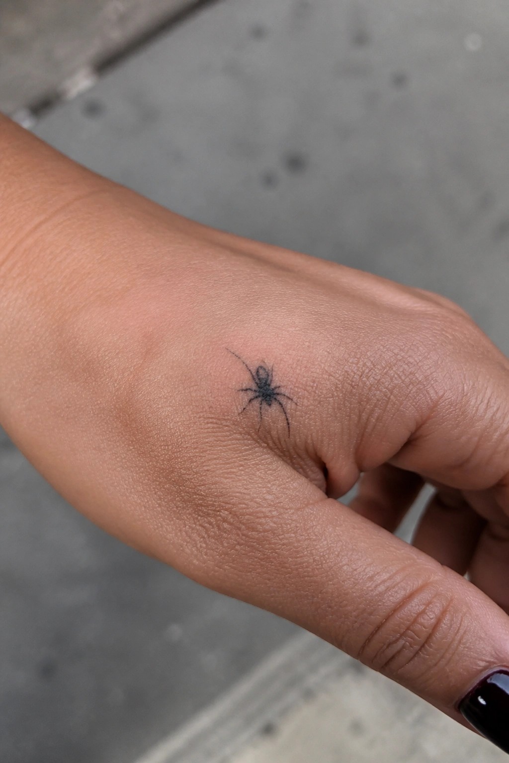 Small black spider tattoo on light brown hand with dark nail polish
