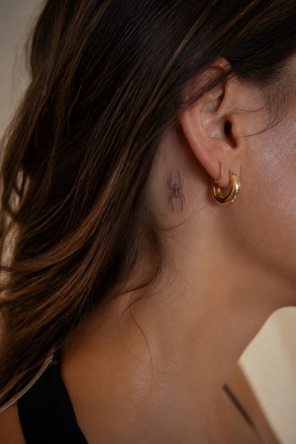 Small spider tattoo behind ear with gold hoop earrings and brown hair